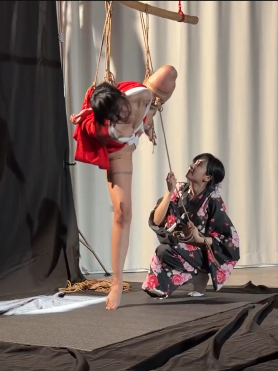 Bunny, kneeling in a black floral kimono, works the rope of a guest model suspended in a red kimono — live, Bangkok, 23.10.26